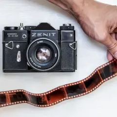 Dwie ręce trzymają kliszę filmową obok czarnego aparatu Zenit TTL. To klasyczna fotografia analogowa.
