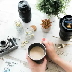 Kawa i sprzęt fotograficzny na biurku, jak w ciemni. Aparat, obiektywy, spinacze i roślina.
