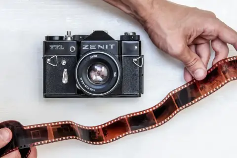 Dwie ręce trzymają kliszę filmową obok czarnego aparatu Zenit TTL. To klasyczna fotografia analogowa.