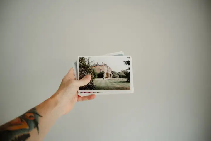 Dłoń trzyma stos zdjęć w formacie 9x13, ukazujących dworek otoczony zielenią.