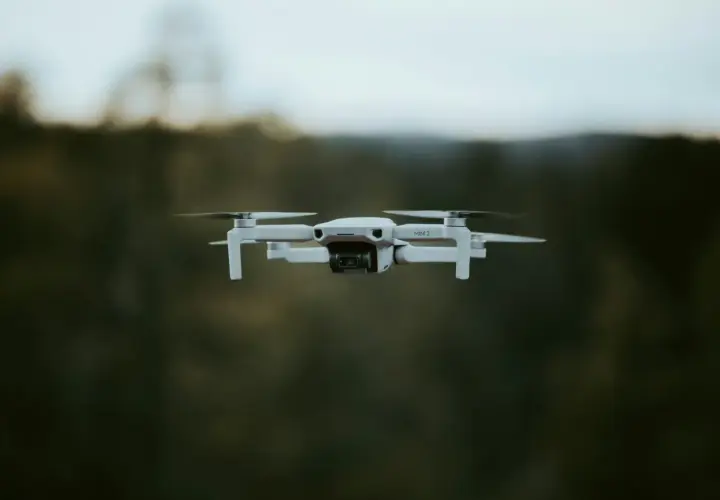 Biały dron DJI Mini 2 w locie, z widocznym obiektywem kamery. Tło to rozmyty las i niebo.