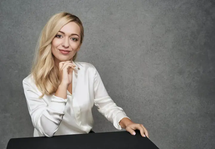 Blondynka w białej satynowej koszuli podczas sesji modowej, z dłonią pod brodą, uśmiecha się do obiektywu.