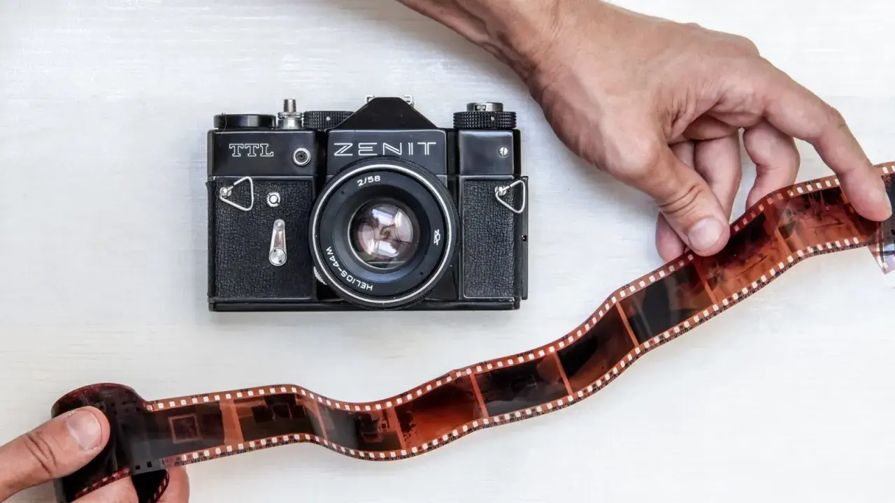 Dwie ręce trzymają kliszę filmową obok czarnego aparatu Zenit TTL. To klasyczna fotografia analogowa.