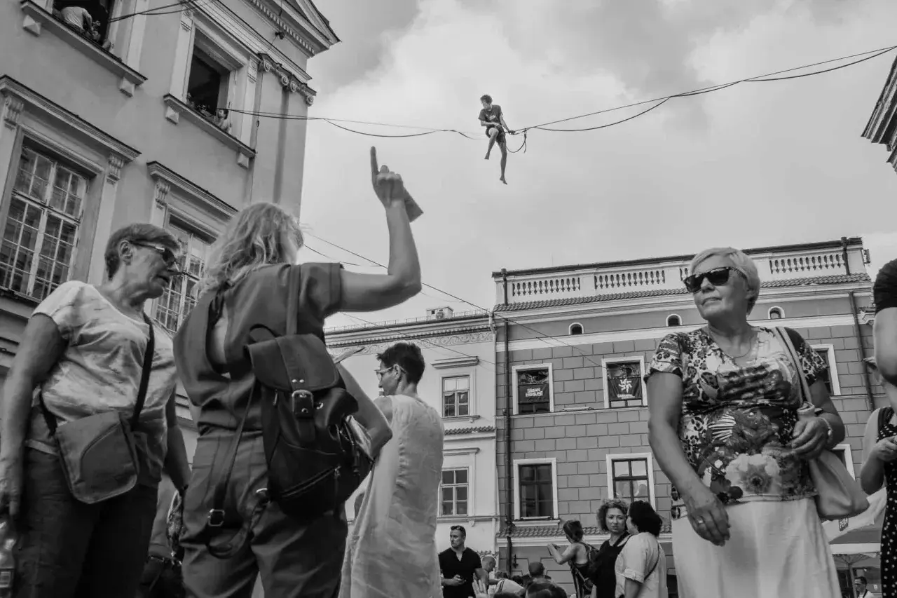 Street photography: widzowie patrzą na akrobatę na linie. Kobieta wskazuje palcem.