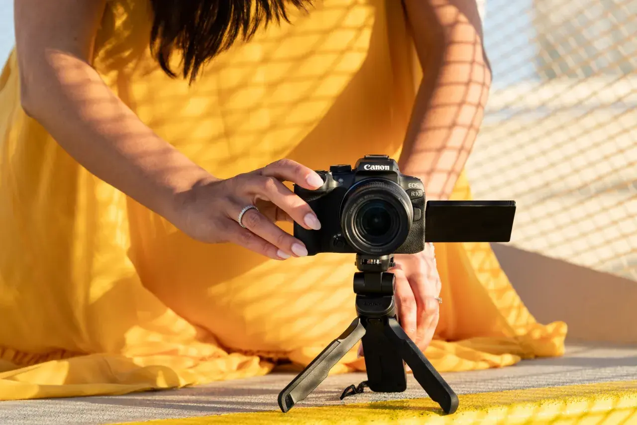 Kobieta w żółtej sukience ustawia aparat Canon EOS R10 na statywie, eksperymentując z trybem manualnym w aparacie.