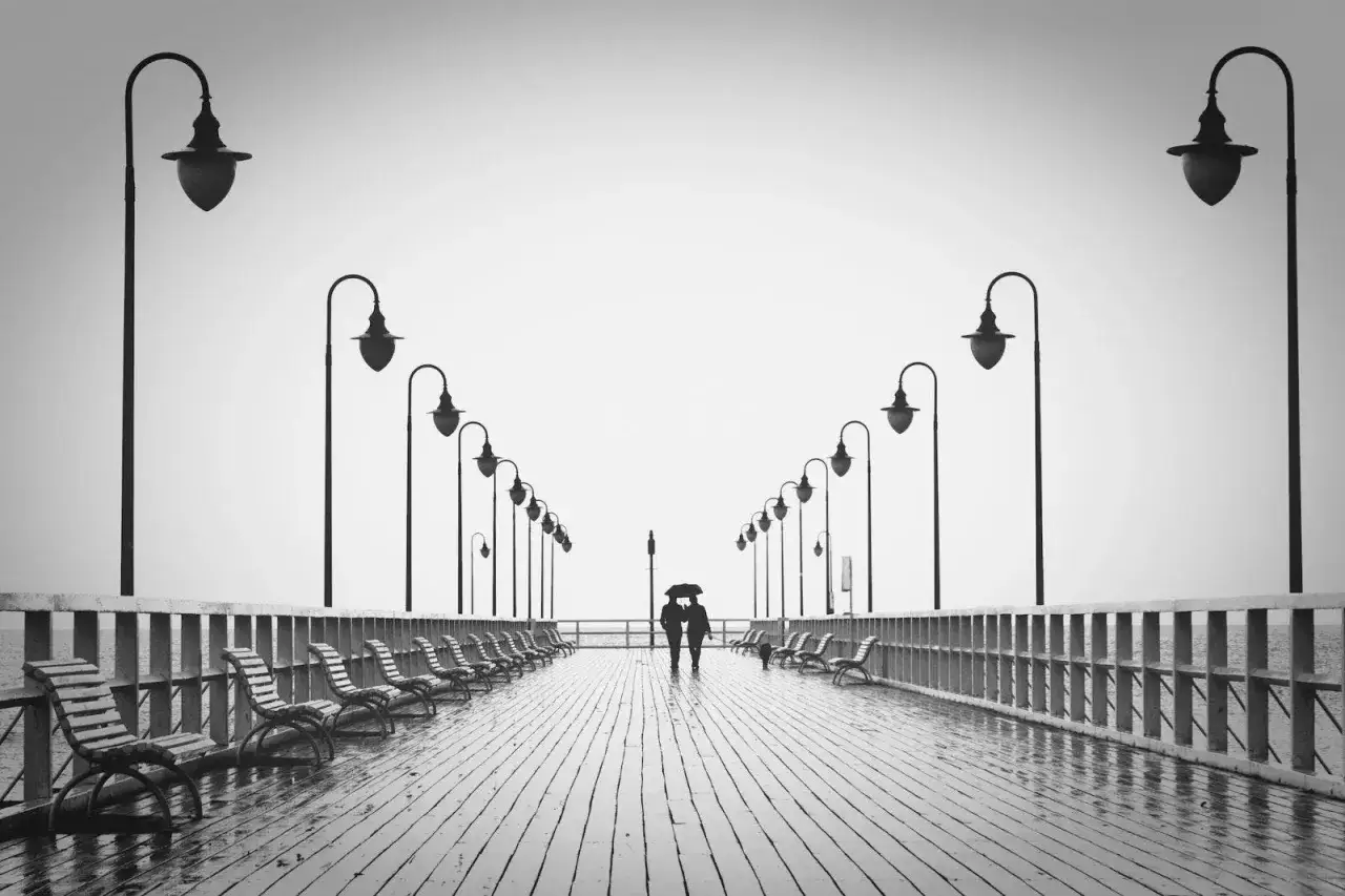 Para spaceruje pod parasolem po moktym molo, otoczona rzędami latarni i pustych ławek. Ustawienia aparatu uchwyciły tę melancholijną scenę.