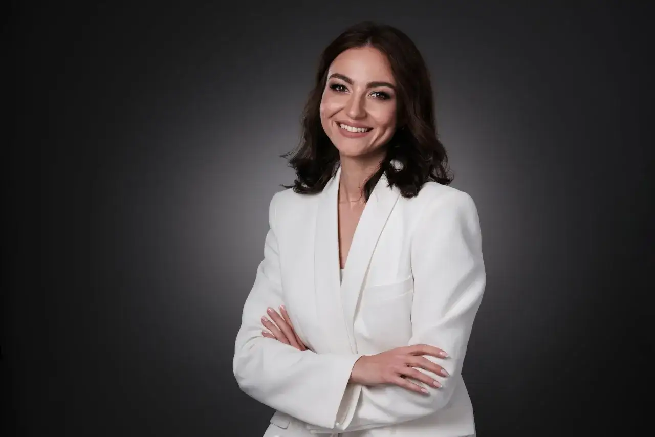 Uśmiechnięta kobieta w białej marynarce, z rękami skrzyżowanymi na piersi, jak po udanym kursie pozowania do zdjęć.